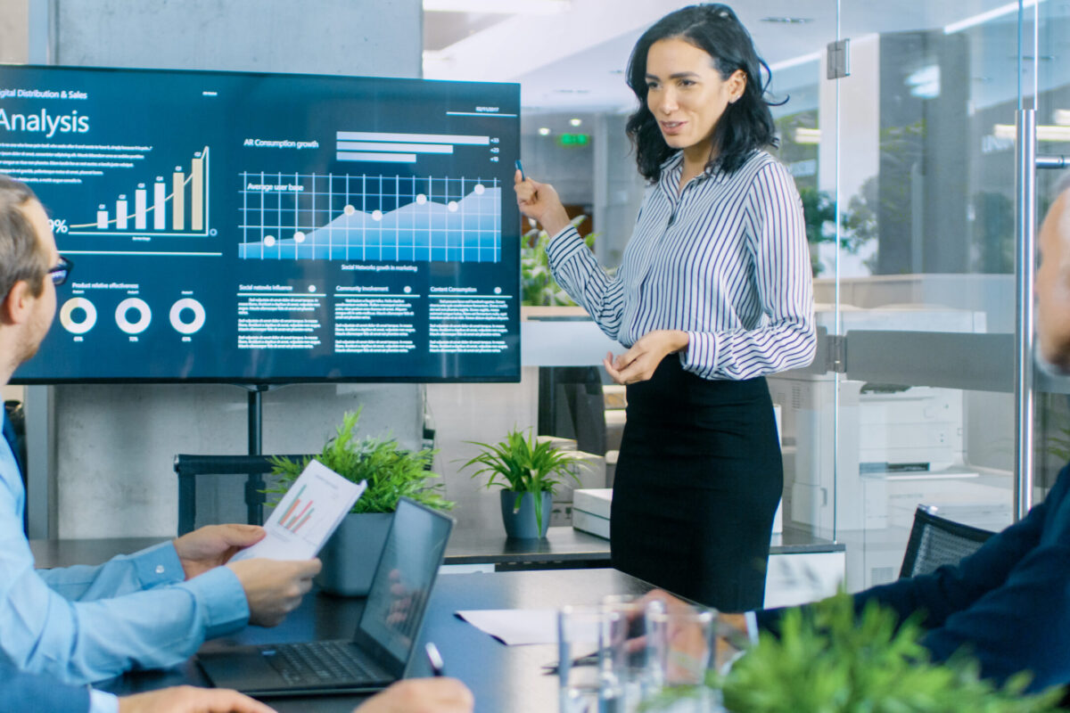 Beautiful Businesswoman Gives Report/ Presentation to Her Business Colleagues in the Conference Room, She Shows Graphics, Pie Charts and Company's Growth on the Wall TV.