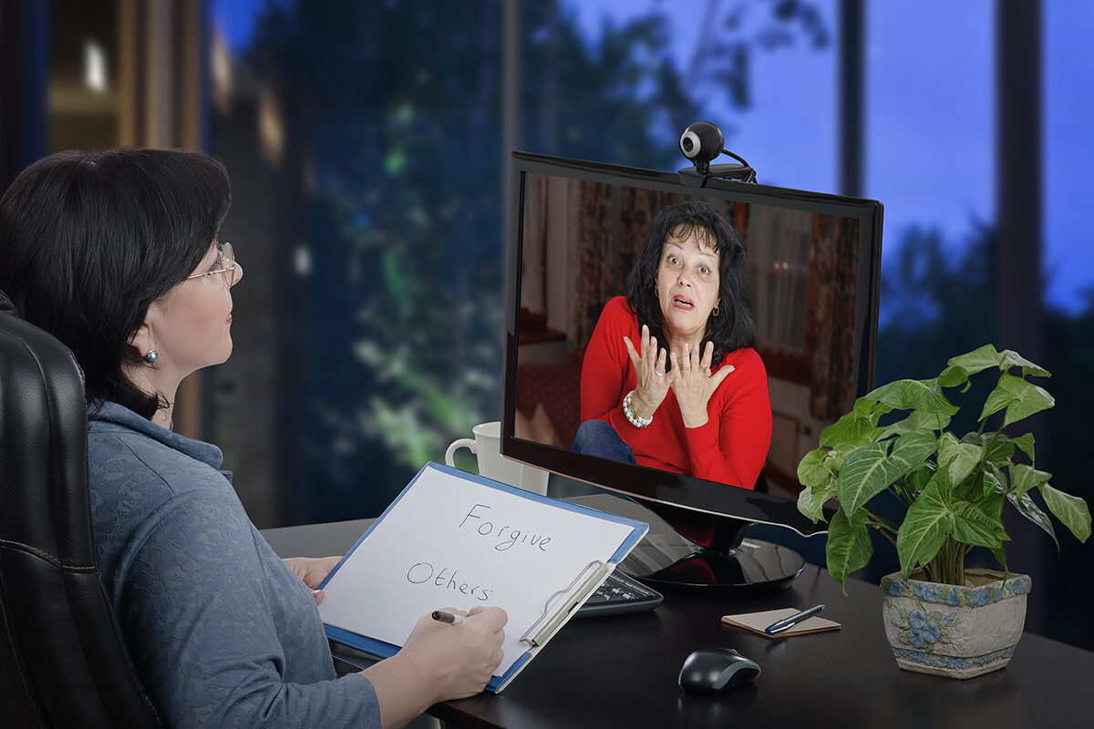 Mature woman in red is on the screen of computer. She explains online her fears and worries to the psychologist woman who sits in the office chair in front of the desktop screen and carefully listens to the patient making some notes in paper pad.