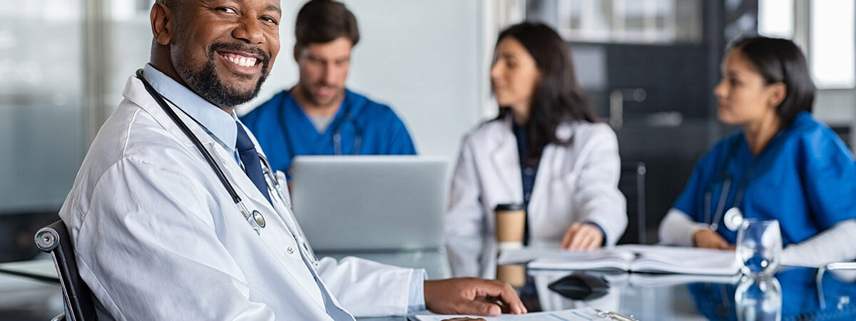 Successful mature doctor at hospital meeting Portrait of cheerful doctor looking at camera and smiling with working clinicians behind. Mature african doctor and medical staff working in hospital. Successful clinic worker in labcoat and stethoscope sitting in conference room with copy space.